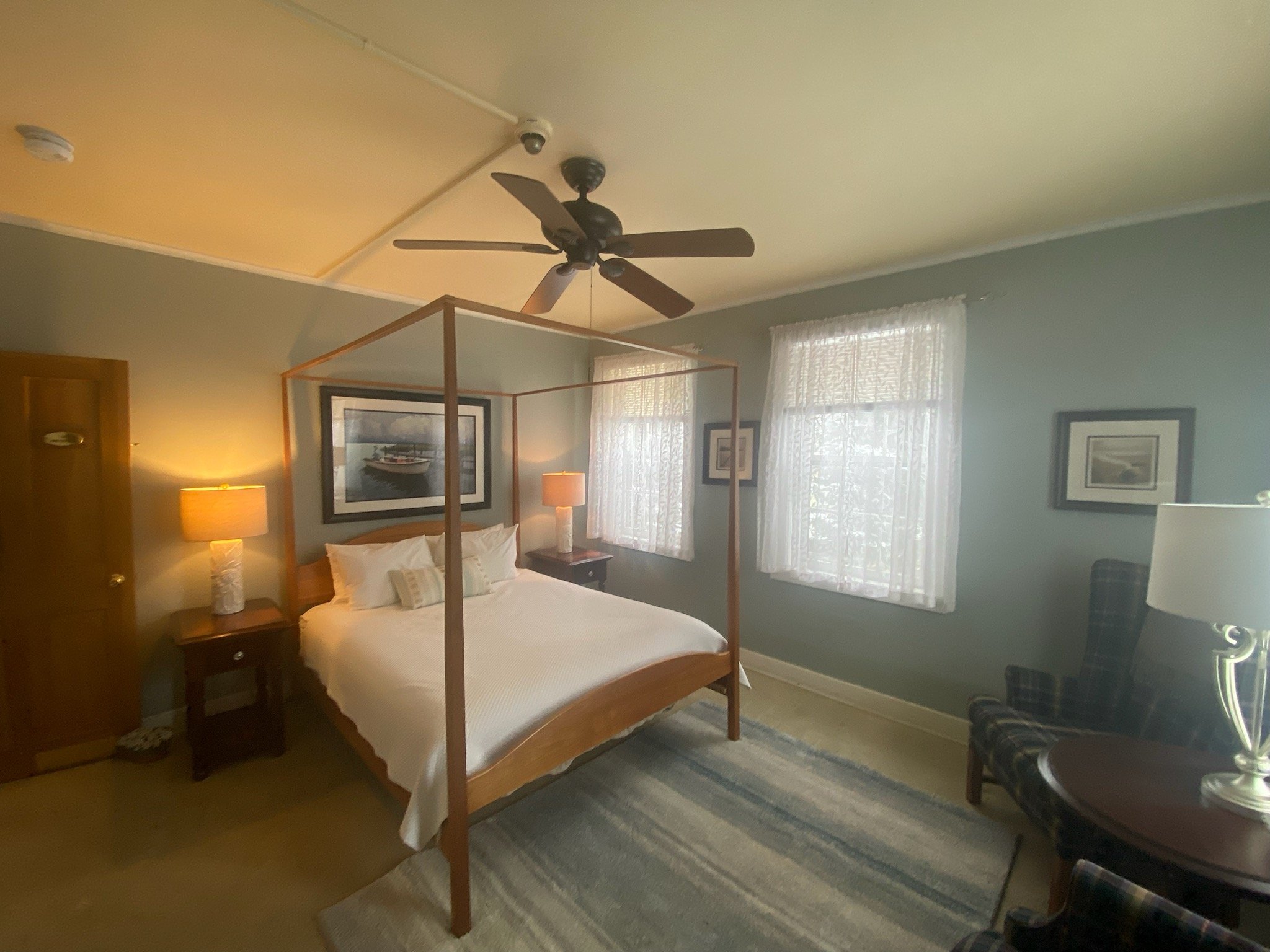 A bedroom with a four-poster bed, two nightstands with lamps, and a ceiling fan. The room has two windows with sheer curtains and a plaid armchair with a side table.