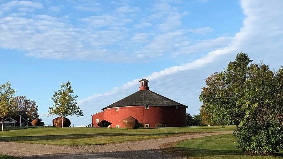 A round red barn with a dark roof and a small tower on top, surrounded by green grass and trees, under a partly cloudy sky.
