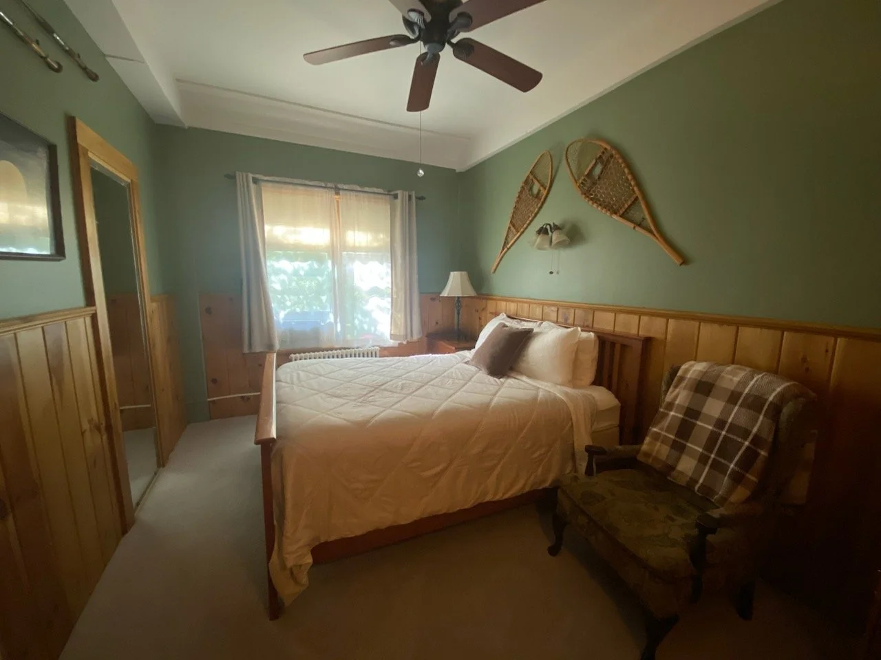A cozy bedroom with wooden furniture including a bed, nightstand, and armchair. There is a window with curtains, a ceiling fan, and vintage decor on the walls.