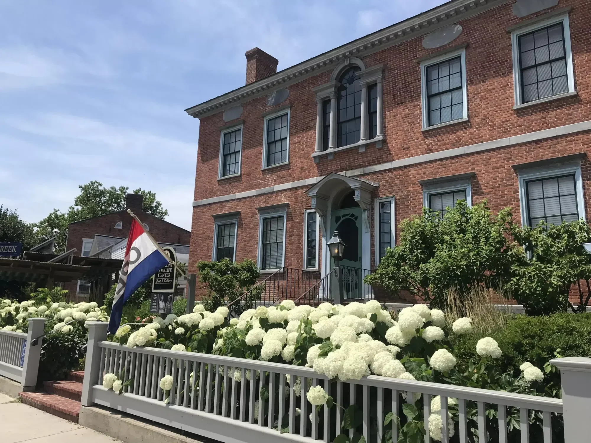 A brick building with multiple windows and a front door with a small awning, surrounded by white hydrangea flowers and some greenery, with a flagpole and a flag in front.
