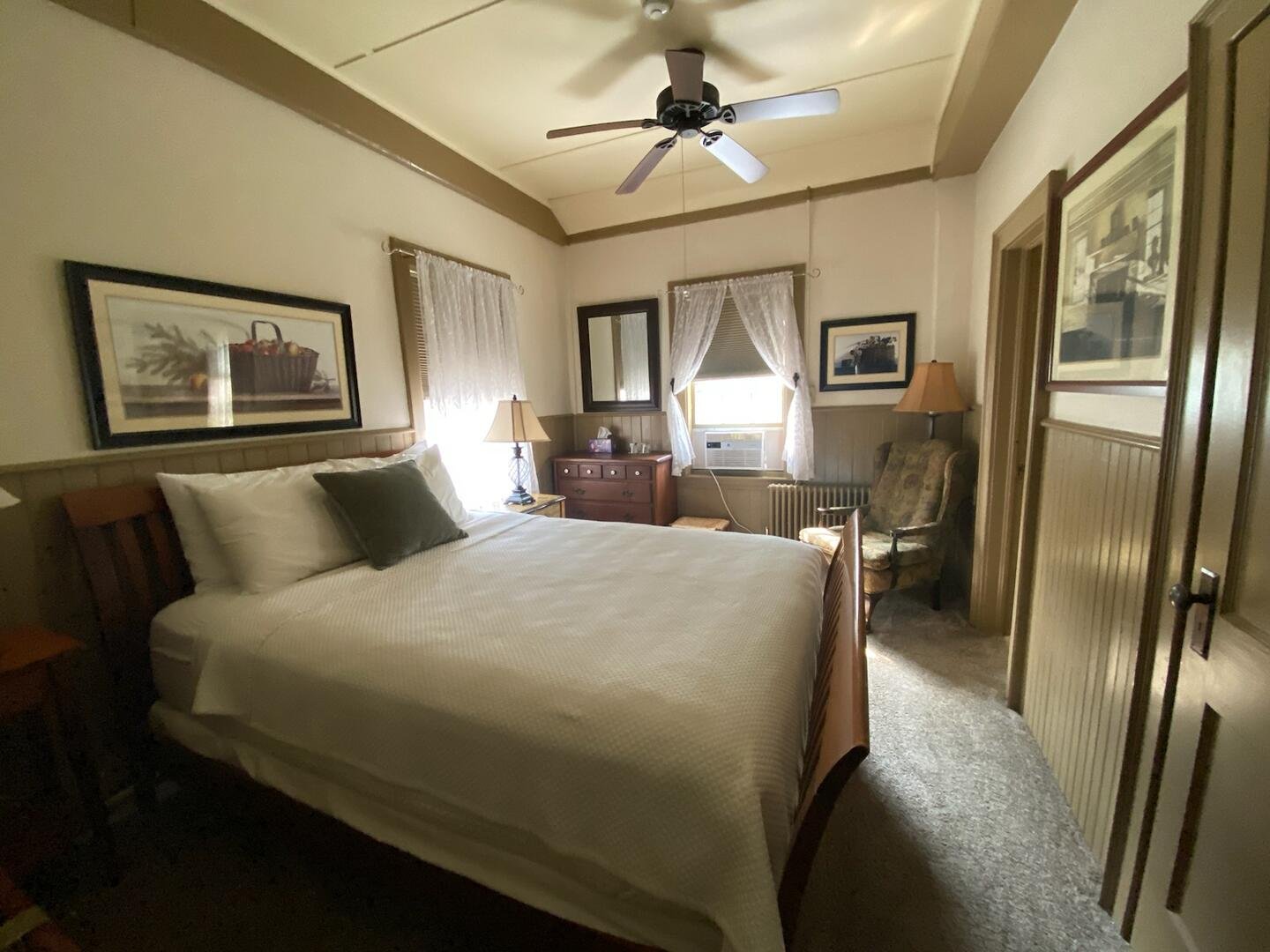 A cozy bedroom with a large bed covered in white bedding, accented by a dark green pillow. The room features two windows with lace curtains, and a ceiling fan. There are bedside lamps, and a small wooden dresser with a mirror on top. An upholstered armchair is next to a radiator near the window. Framed artwork decorates the walls.