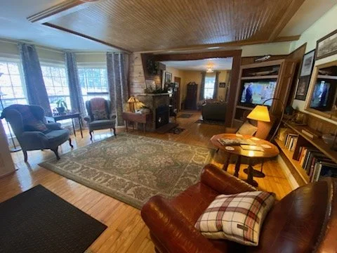 Living room with wooden floors, large window with curtains, comfortable armchairs, a rug, a fireplace, and a TV. There are bookshelves and a side table with a lamp.