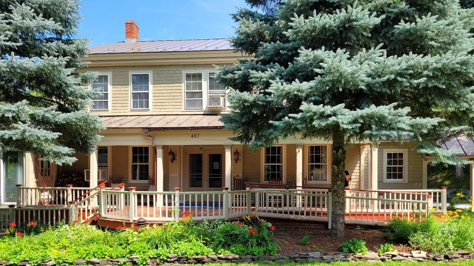A large, two-story yellow house with a brown roof, surrounded by lush green trees and plants. The house has a porch with a railing and several windows, with the house number 457 displayed above the doors.