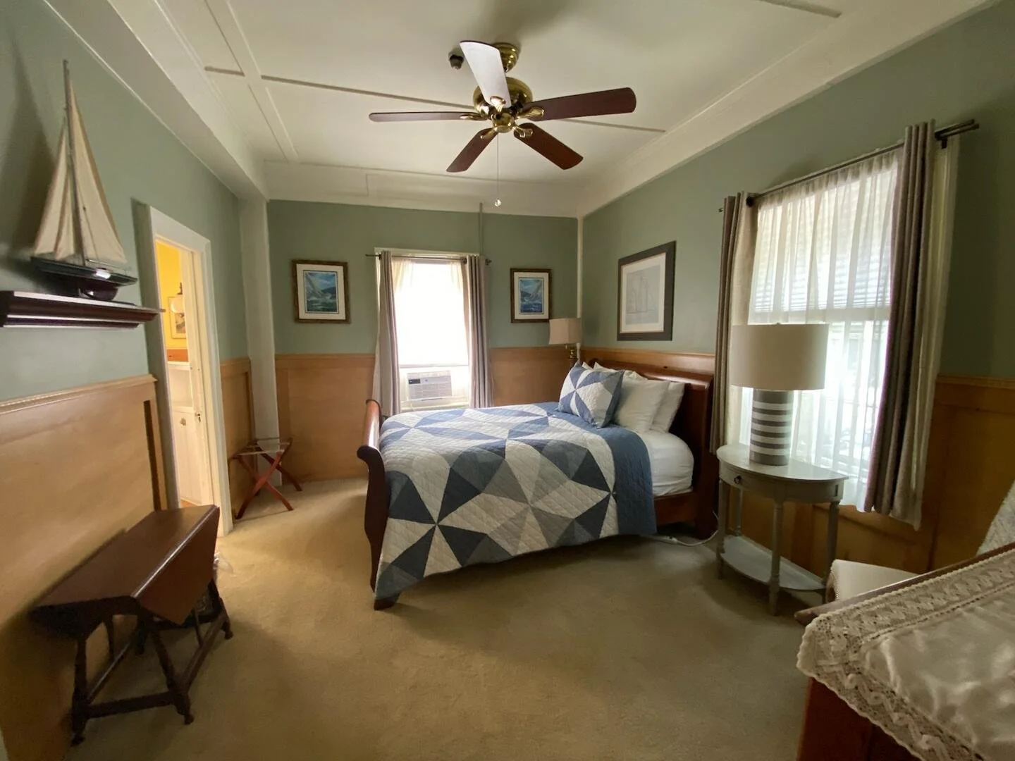 A cozy bedroom with a green wall, wooden accents, a ceiling fan, a bed with a blue and white quilt, and natural light coming through two windows with curtains.