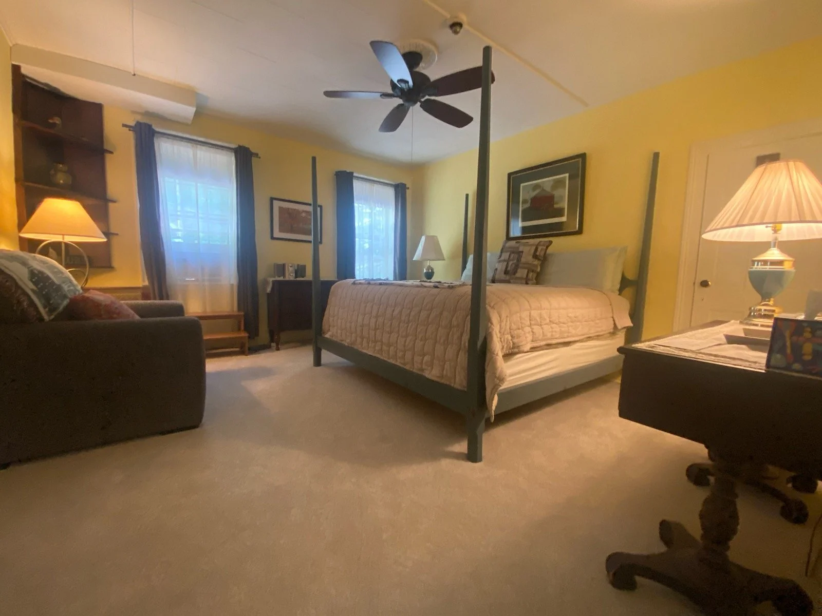 A bedroom with a large bed, decorated with pillows and a quilt, a ceiling fan, a small dresser and nightstand with a lamp, a dark-colored sofa with pillows, and windows with curtains, warm lighting, and framed artwork on the walls.