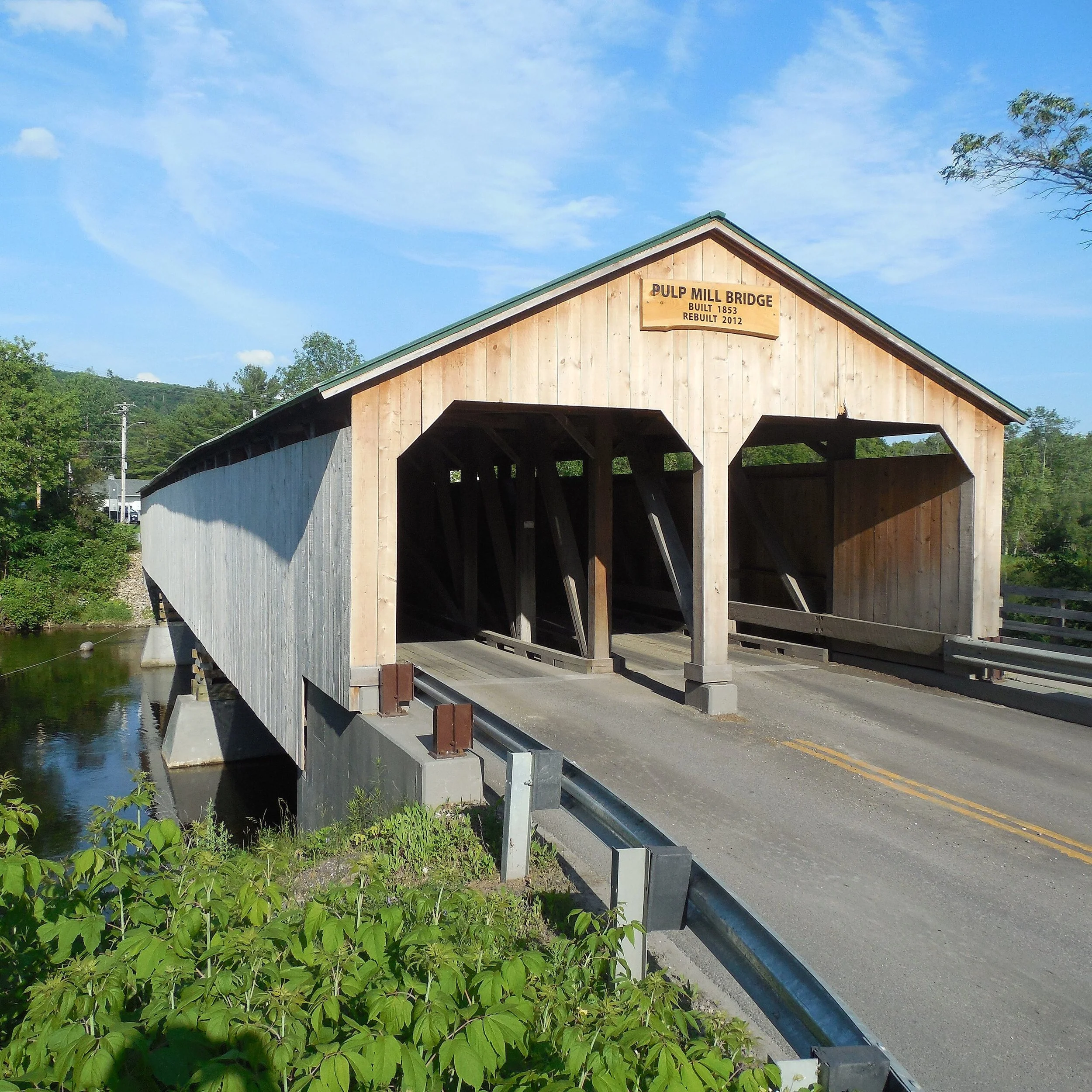 Covered wooden bridge over a river, labeled 'Pulp Mill Bridge', built in 1853, rebuilt in 2012, under a blue sky with trees on the sides.