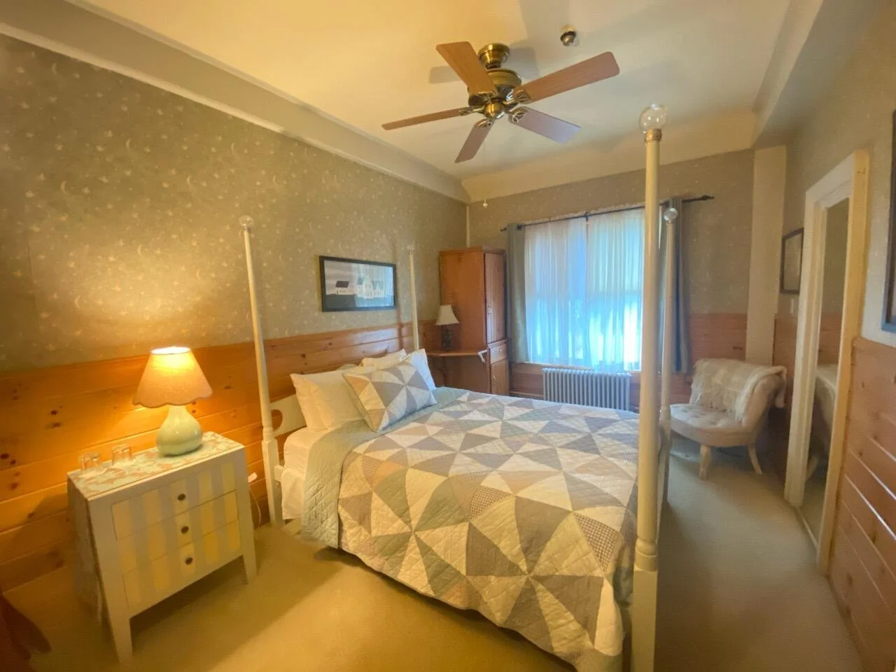 A cozy bedroom with a four-poster bed, a patchwork quilt, a bedside table with a lamp, a window with curtains, a chair, and wooden wall paneling.