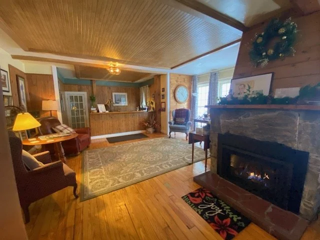Living room with wood-paneled walls and ceiling, a stone fireplace with a holiday wreath on the mantel, a large patterned area rug, a leather sofa, armchairs, a side table with a yellow lamp, and a reception desk in the background near a door with gl