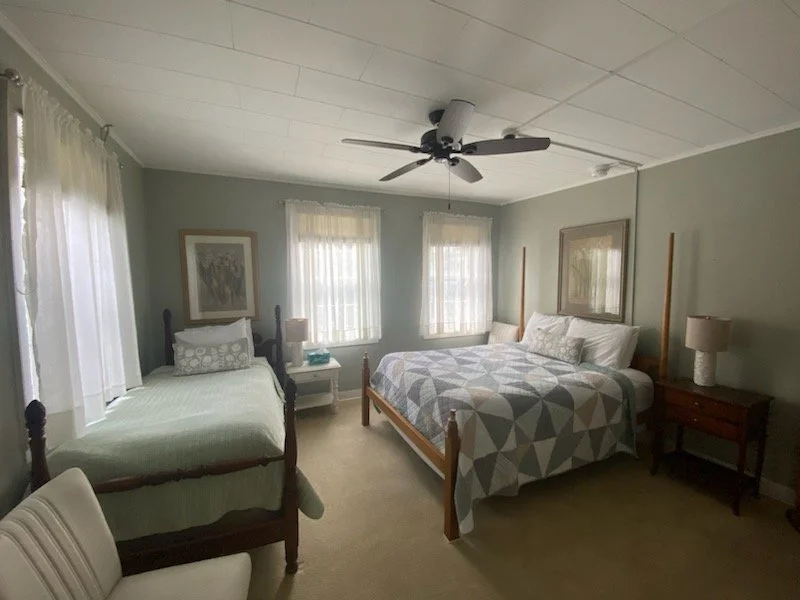 A bedroom with a twin bed on the left and a larger bed on the right, both with pillows and bedding. There are two windows with sheer curtains, a ceiling fan, and two nightstands with lamps.