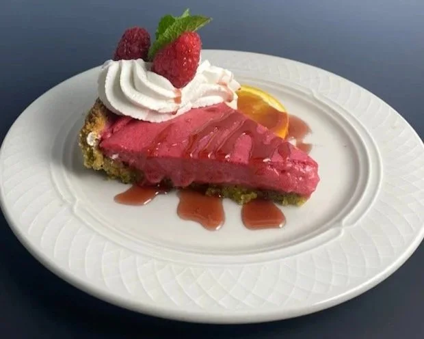 Slice of raspberry pie with whipped cream, raspberries, a mint leaf, and a lemon wedge on a white plate.