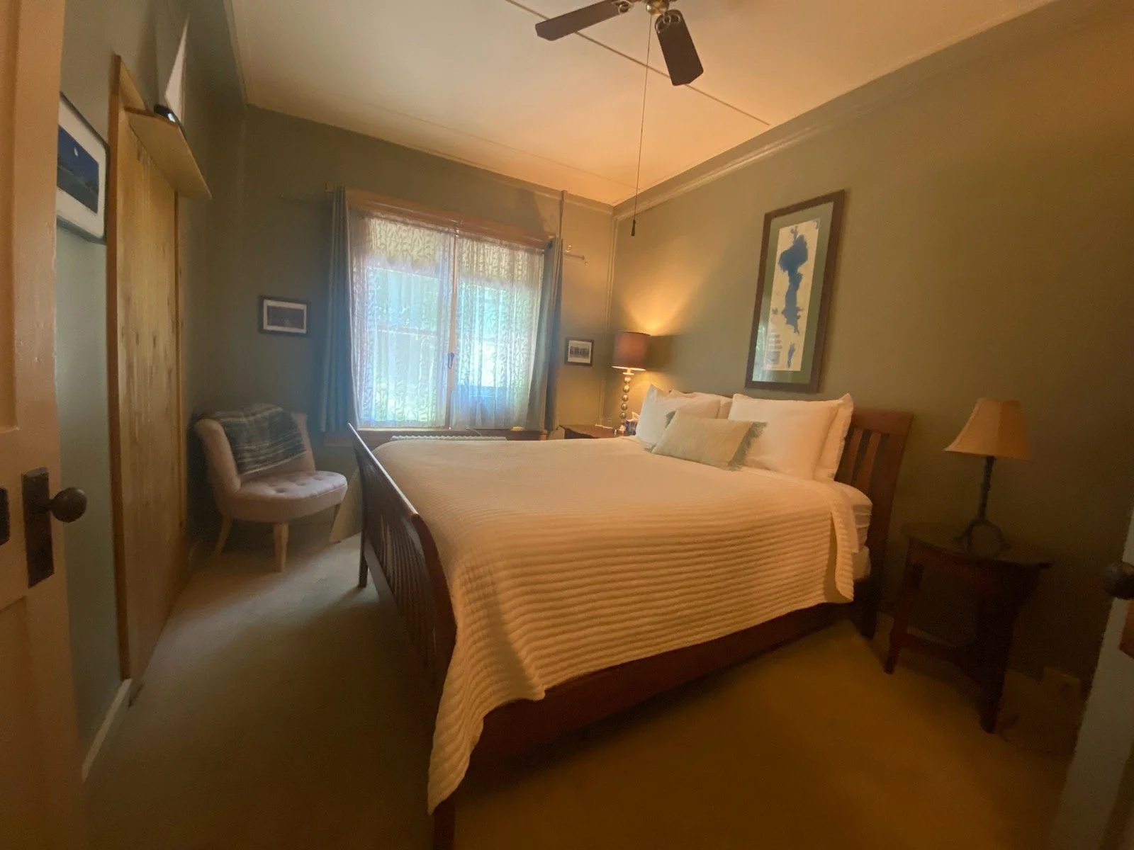 A cozy bedroom with a queen-sized bed, a wooden headboard, and a patterned bedspread. There is a window with blue curtains, a small armchair, a side table with a lamp, a framed picture of a sailboat, and a ceiling fan. The walls are painted light green.