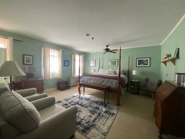 A bedroom with green walls, a four-poster bed, a ceiling fan, a window air conditioner, a leather couch, a patterned rug, a wooden dresser, a chair, and framed pictures on the walls.