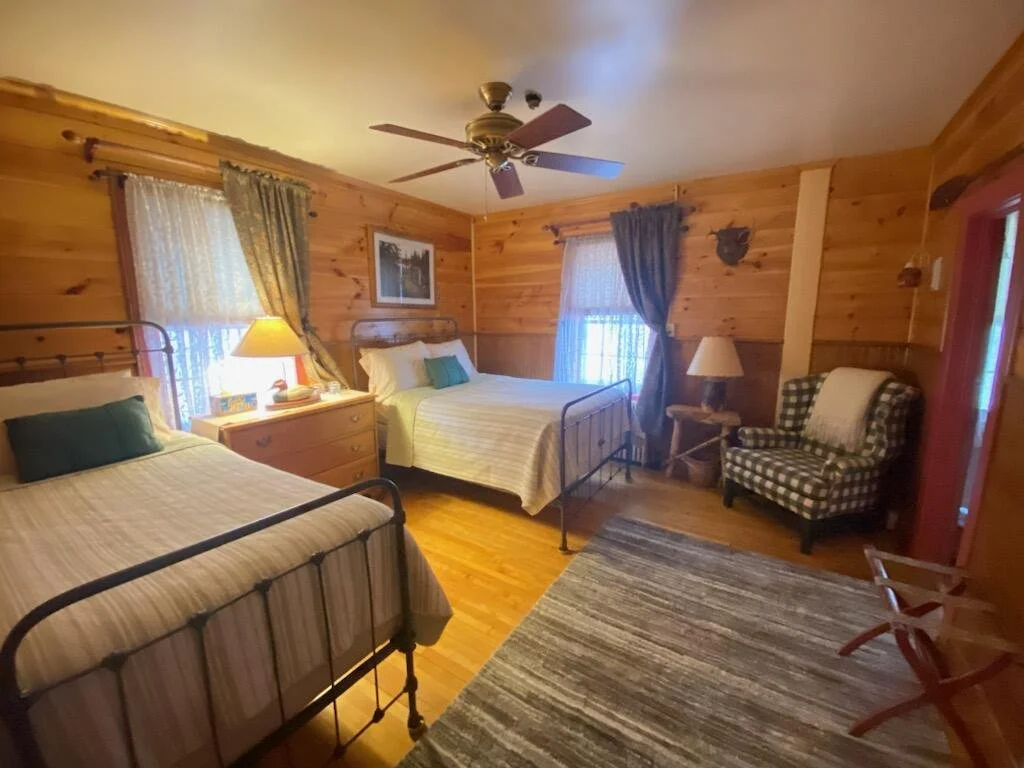 Room with two beds, wooden walls, ceiling fan, window with curtains, armchair, dresser with lamp, and a rug.