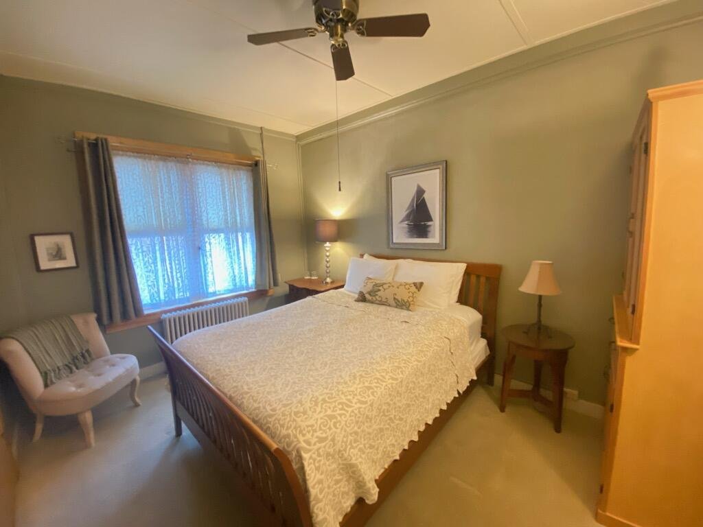 A cozy bedroom with a queen-sized bed, a wooden headboard, and a patterned bedspread. There is a window with blue curtains, a small armchair, a side table with a lamp, a framed picture of a sailboat, and a ceiling fan. The walls are painted light green.