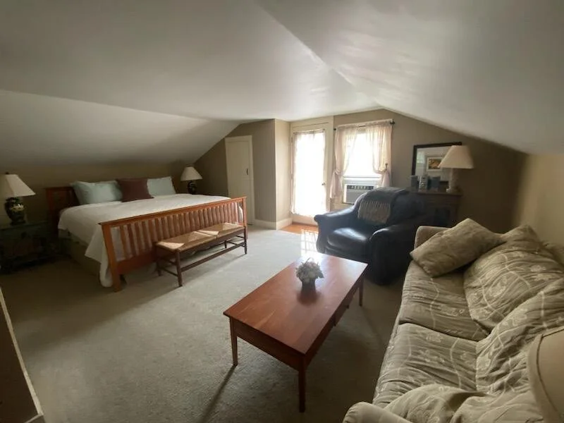 A cozy bedroom with a sloped ceiling, featuring a bed with pillows, two nightstands with lamps, a bench at the foot of the bed, a blue armchair, a black sofa, a wooden coffee table with a small flower arrangement, and a window with curtains letting in natural light.