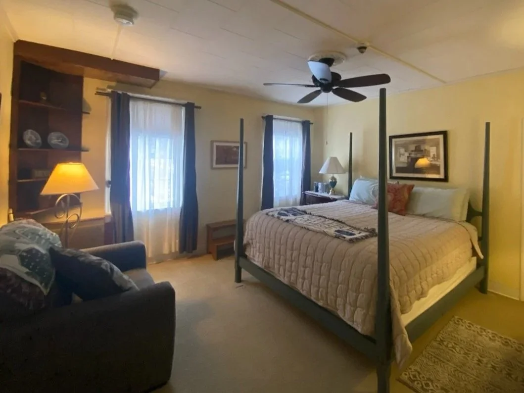 A bedroom with a large bed, decorated with pillows and a quilt, a ceiling fan, a small dresser and nightstand with a lamp, a dark-colored sofa with pillows, and windows with curtains, warm lighting, and framed artwork on the walls.