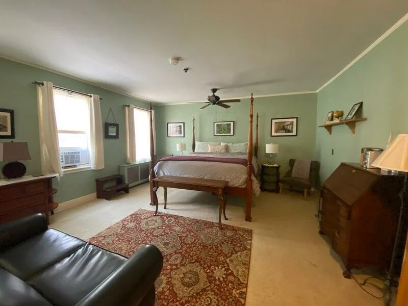 A bedroom with green walls, a four-poster bed, a ceiling fan, a window air conditioner, a leather couch, a patterned rug, a wooden dresser, a chair, and framed pictures on the walls.