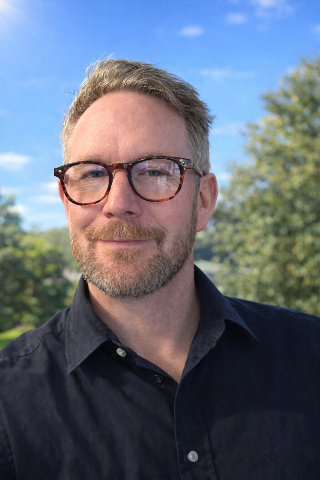 An experienced, reliable, sensitive, life coach & mentor with glasses and a beard smiling outdoors with trees and a blue sky in the background.