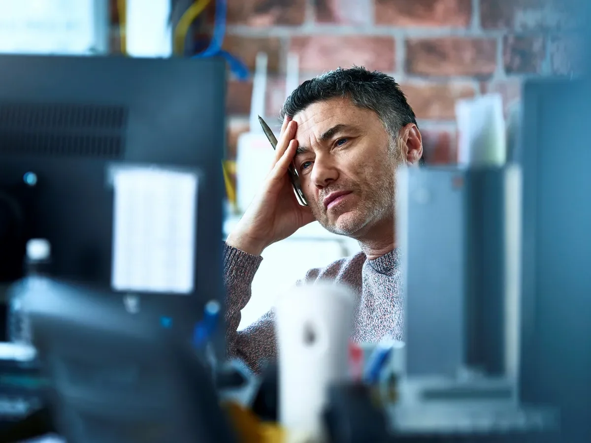 A man sitting at a desk with computer monitors, holding his forehead and looking stressed or frustrated, in a workspace with a brick wall background.