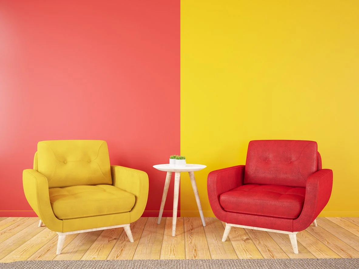 A yellow armchair and a red armchair are separated by a small white round table with a small plant on top. The background has a split-colored wall, pink on the left and yellow on the right, and a wooden floor.