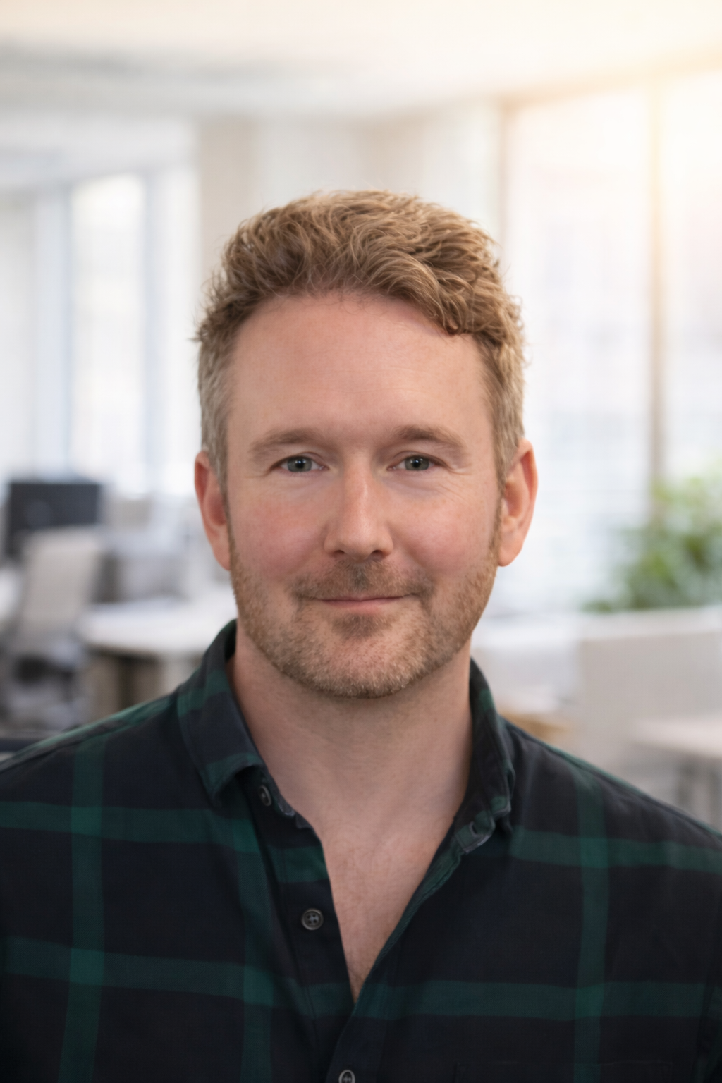 Headshot of an experienced life coach and mentor with short hair, a light beard, wearing a dark green and black plaid shirt, smiling in an office setting with bright natural light.