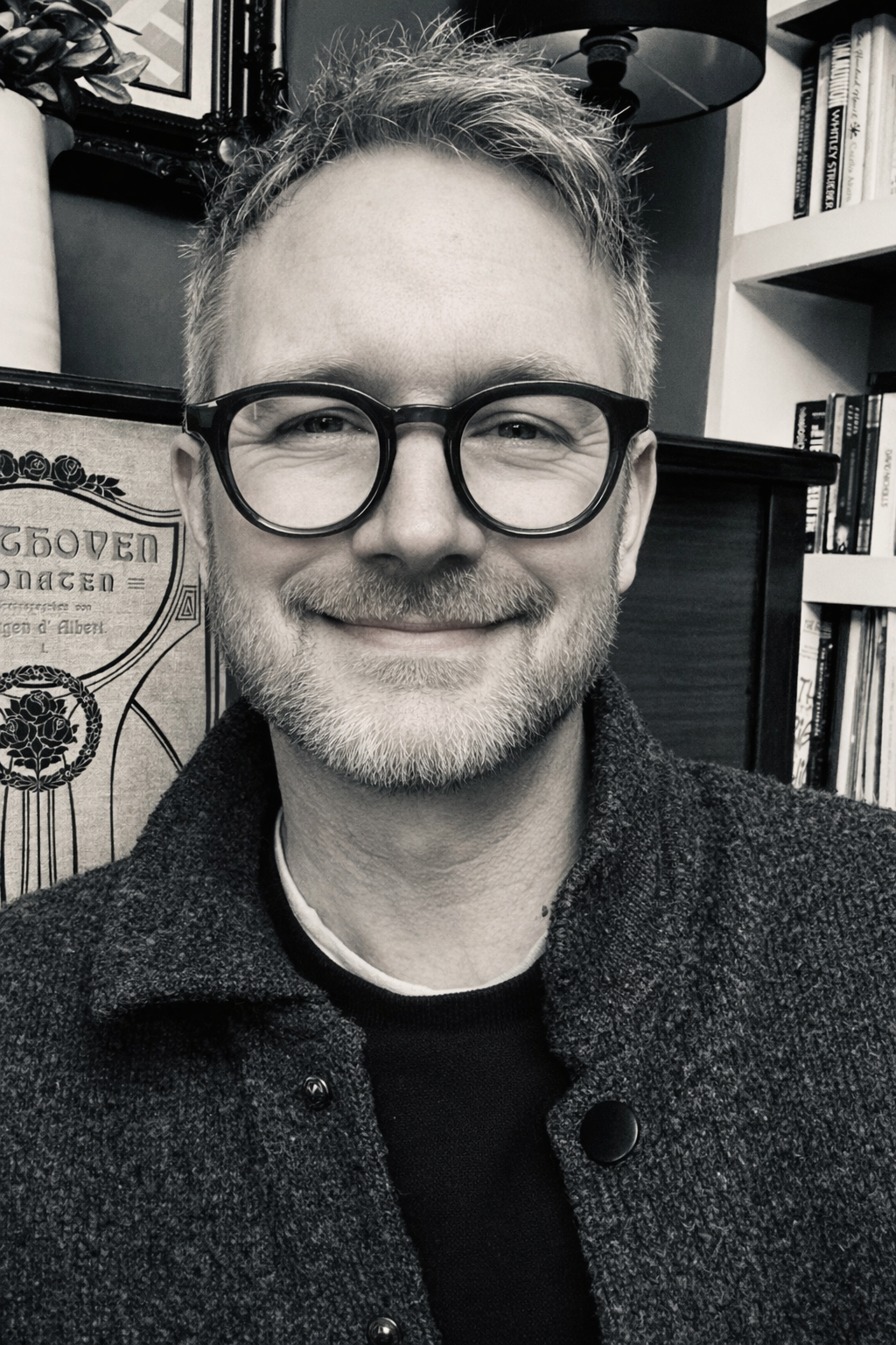 A smiling life coach and mentor with glasses and facial hair, wearing a textured coat, in a room with shelves of books and framed art.