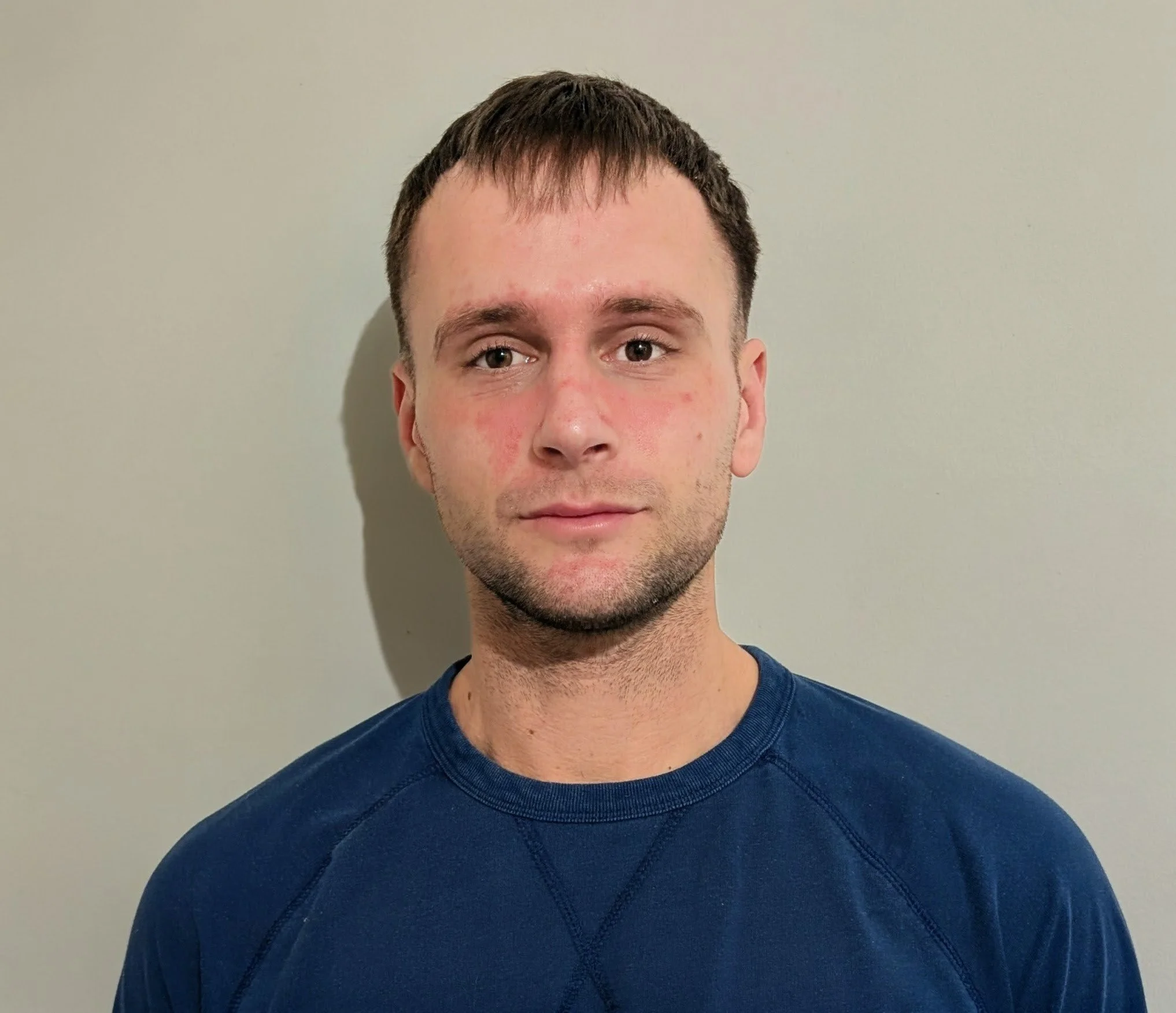 A portrait of a young man with short, dark hair, wearing a navy blue sweatshirt, standing against a plain light-colored wall.