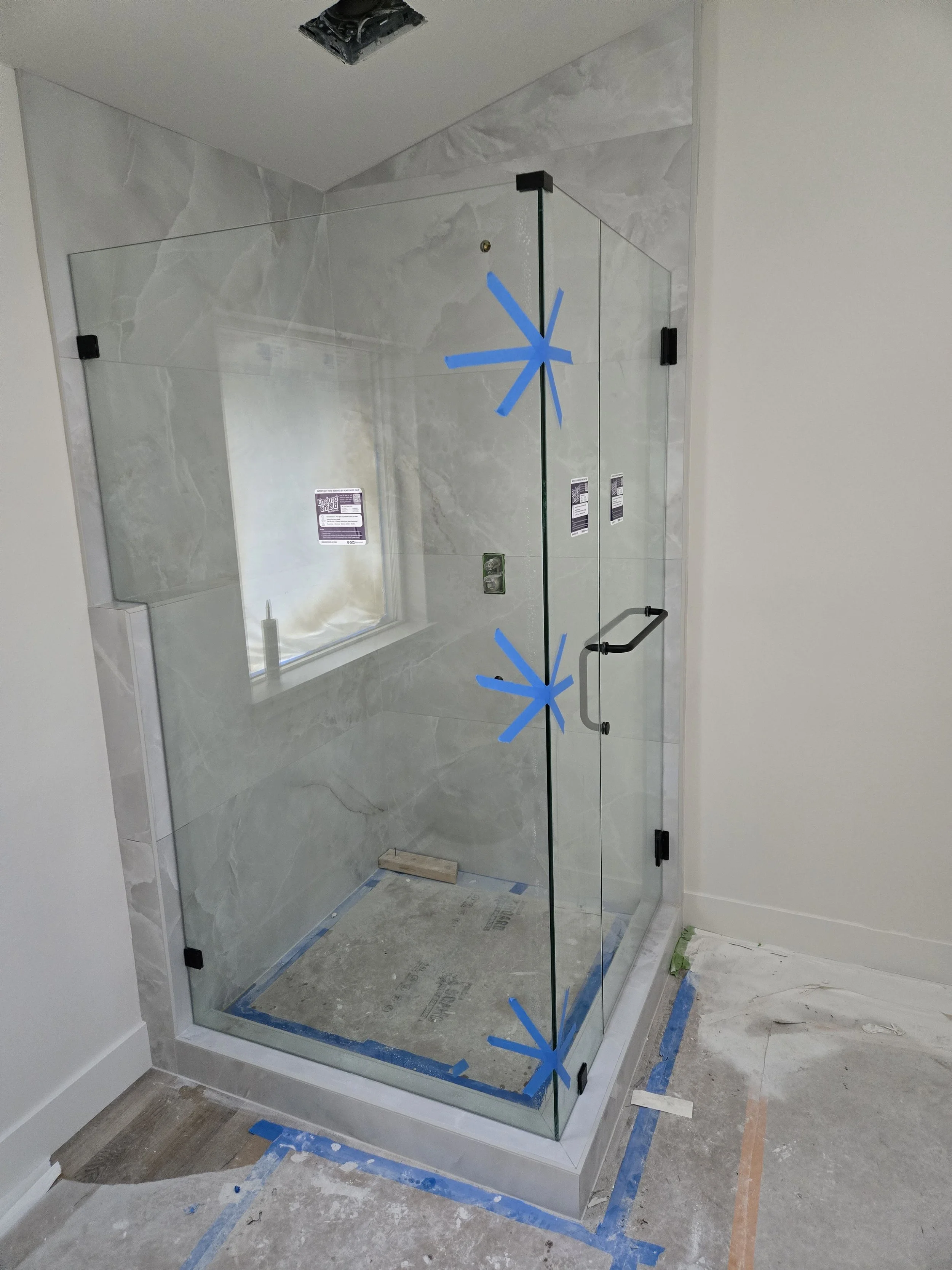 Glass shower enclosure under installation with blue painter's tape markings on the glass. The shower has a marble or tile interior, a metal handle on the door, and construction supplies on the floor.