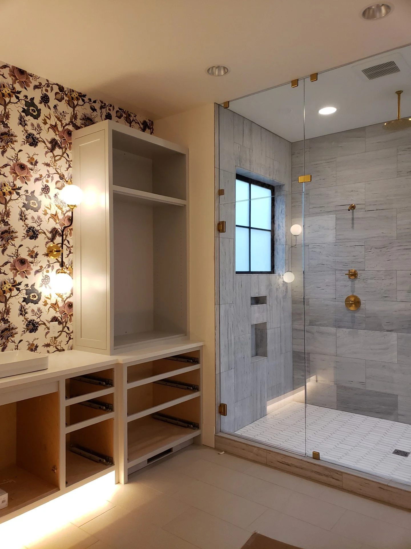A bathroom with a glass shower enclosure, gray marble tiles, a window, a floral wallpapered wall, a white vanity with open drawers, and updated lighting fixtures.
