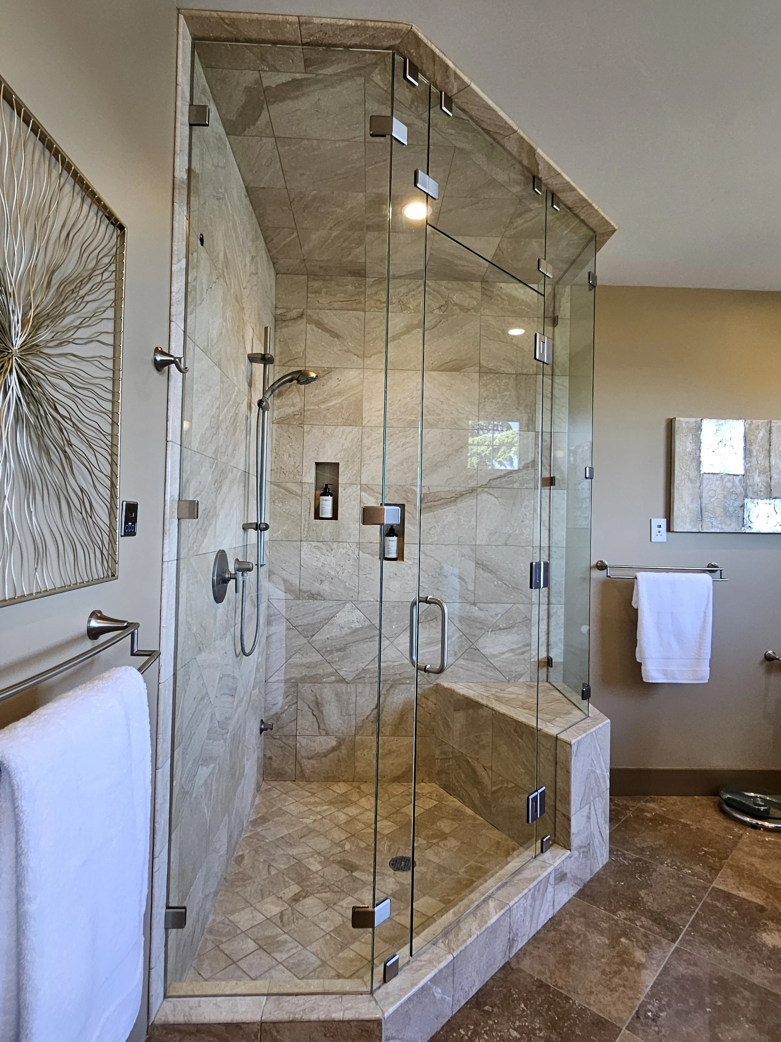 A glass-enclosed walk-in shower with beige marble tiles, a built-in bench, and shower shelves with bottles. Two white towels are hung on towel bars, and a decorative metal wall art piece is on the adjacent wall. The bathroom has a tiled floor and beige painted walls.