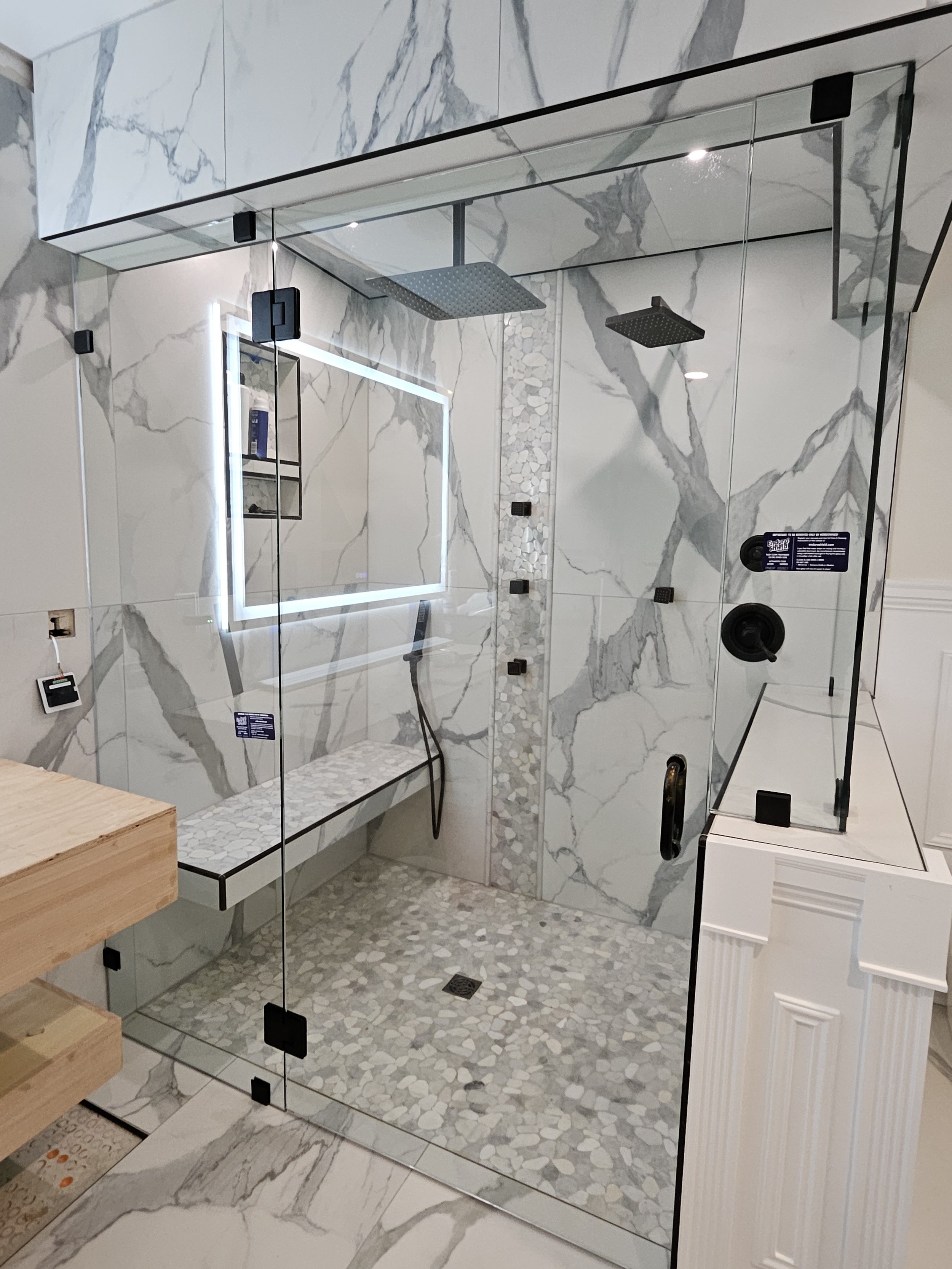 Modern walk-in shower with glass enclosure, marble walls, pebble floor, built-in bench, rainfall and handheld showerheads, and a large illuminated mirror.