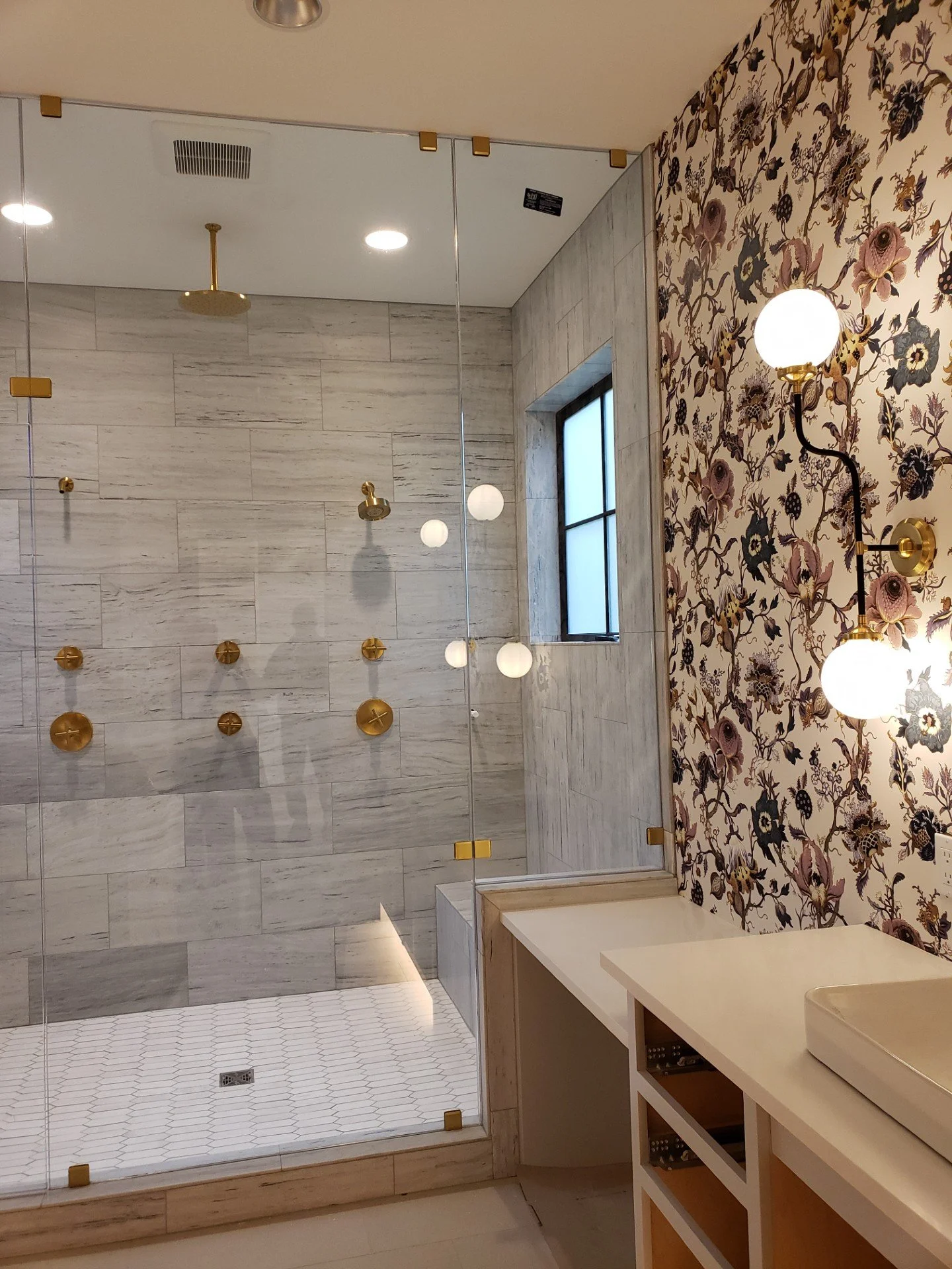 Bathroom with a walk-in shower featuring glass walls, gold fixtures, and a window, decorated with floral wallpaper on the right wall and a white vanity with a sink on the right side.