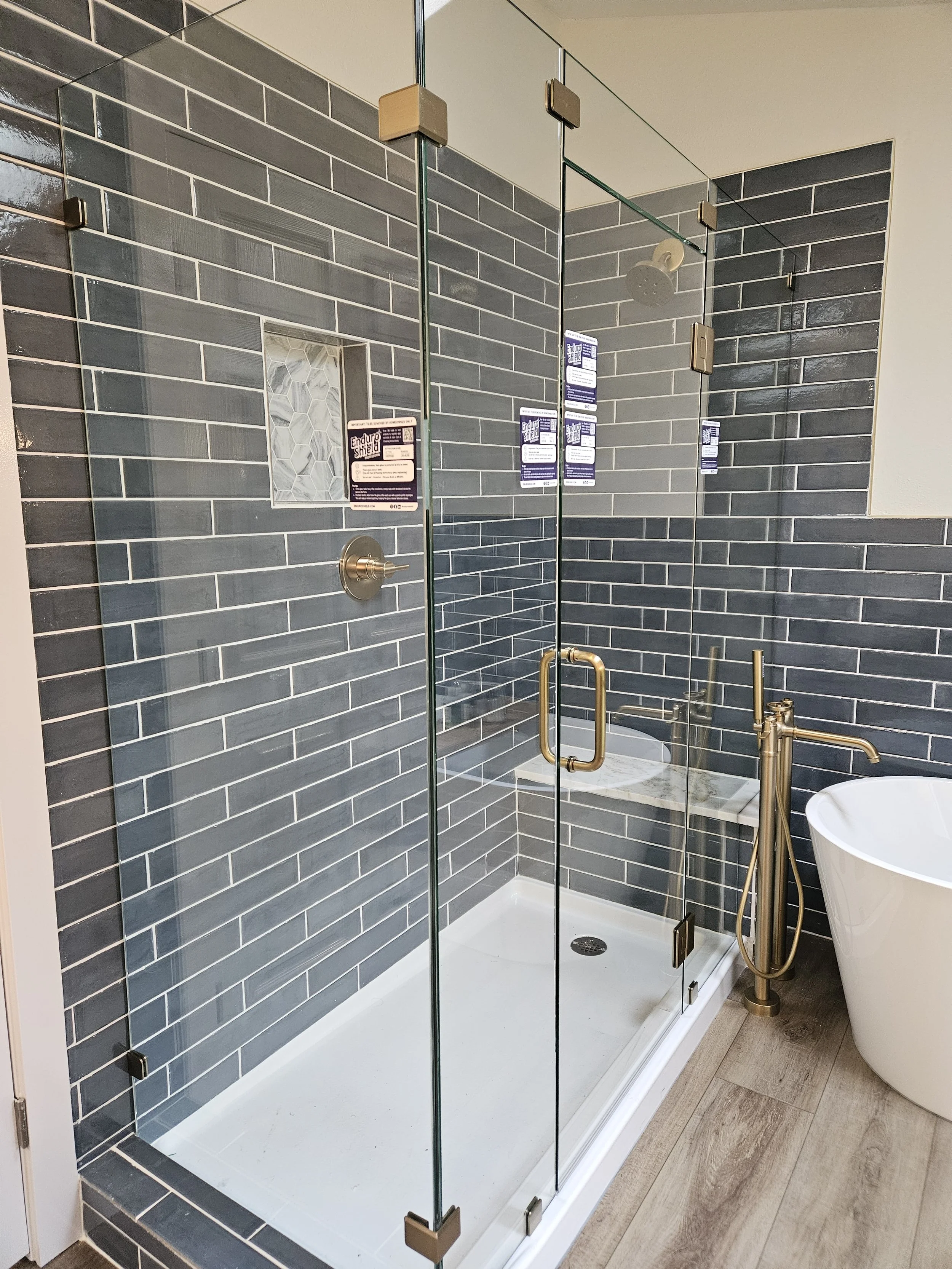 Modern bathroom with a glass shower enclosure featuring dark gray subway tile walls and a built-in niche with a hexagon tile accent. There is a brushed gold showerhead, handle, and a gold floor-standing faucet next to a white freestanding bathtub with a wood-look floor.