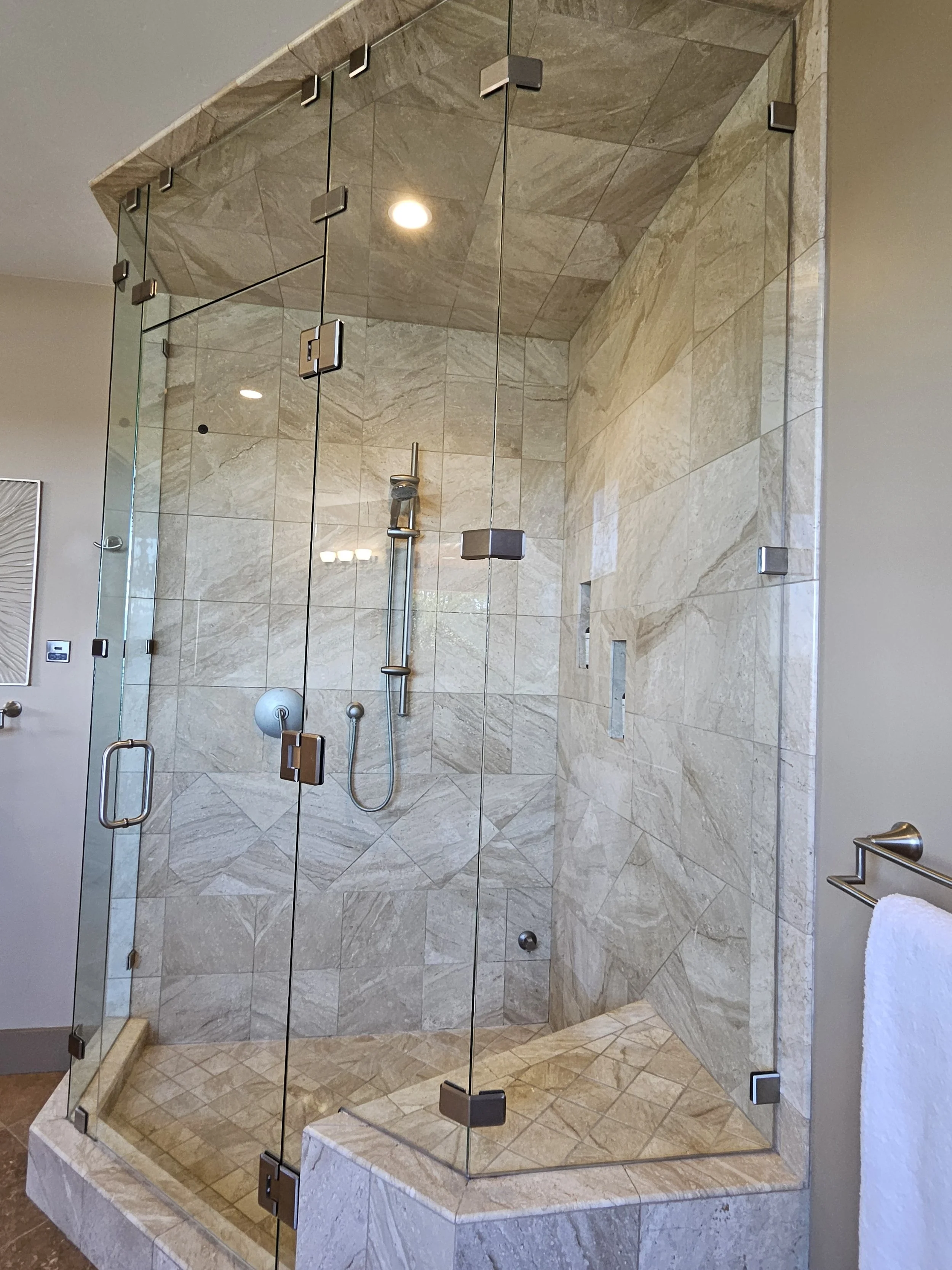 A glass-enclosed shower with beige marble tiles on the walls and floor, a rainfall showerhead, a handheld shower wand, and built-in wall niches for toiletries.