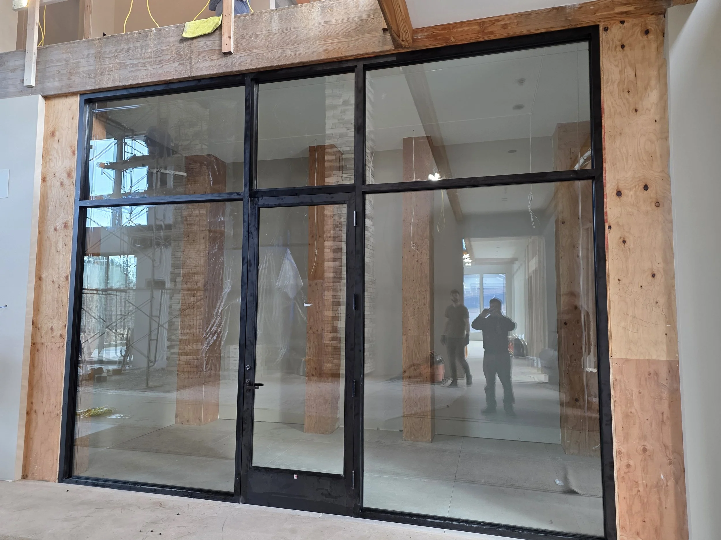 A large glass storefront with black metal framing and a door, under construction inside a building, with two workers and scaffolding visible through the glass.