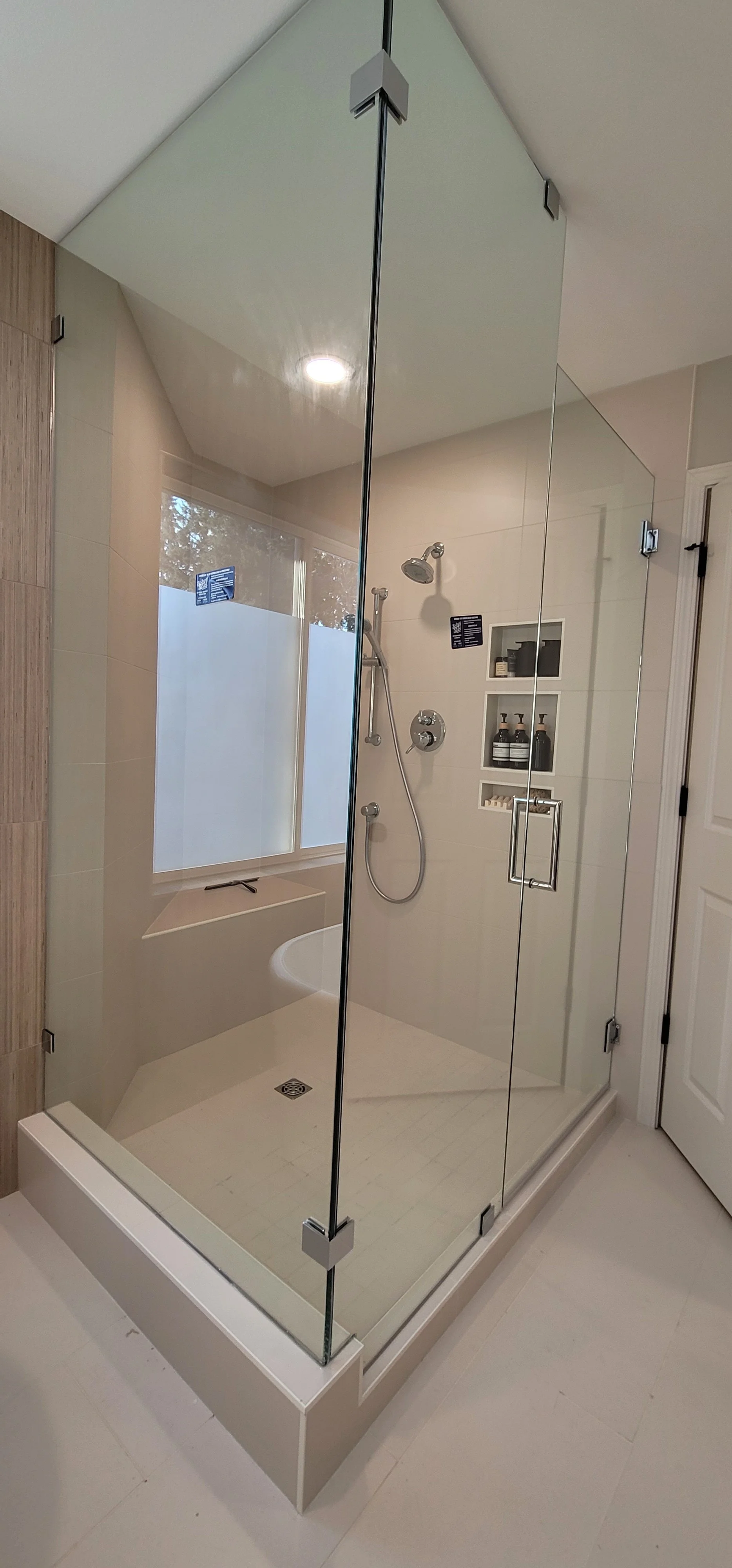 A modern glass shower enclosure in a bathroom with a built-in bench, shower fixtures, and wall niches for toiletries, with a frosted window and light-colored tiles.