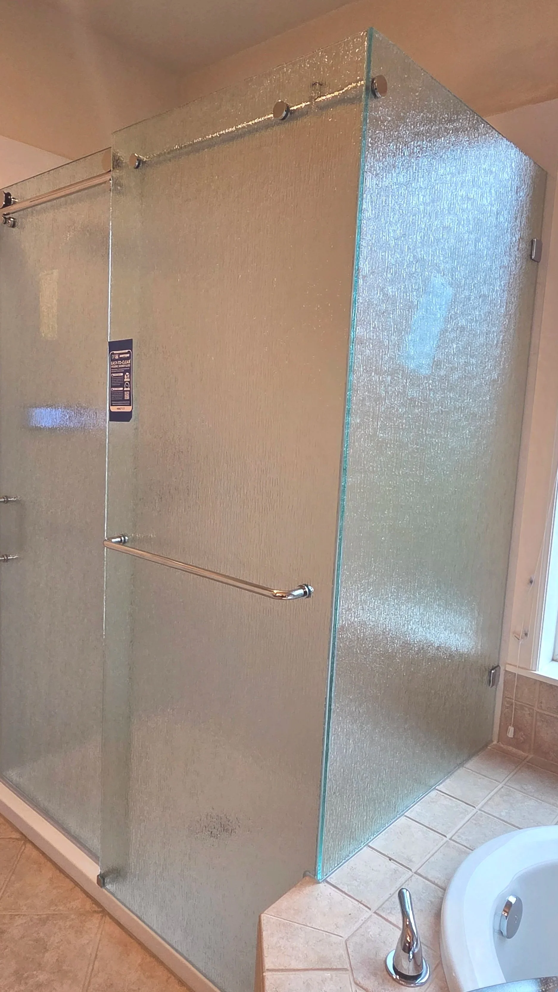 A glass shower enclosure with textured glass panels, metal hardware, and a towel bar inside a bathroom with tiled flooring and a corner bathtub.