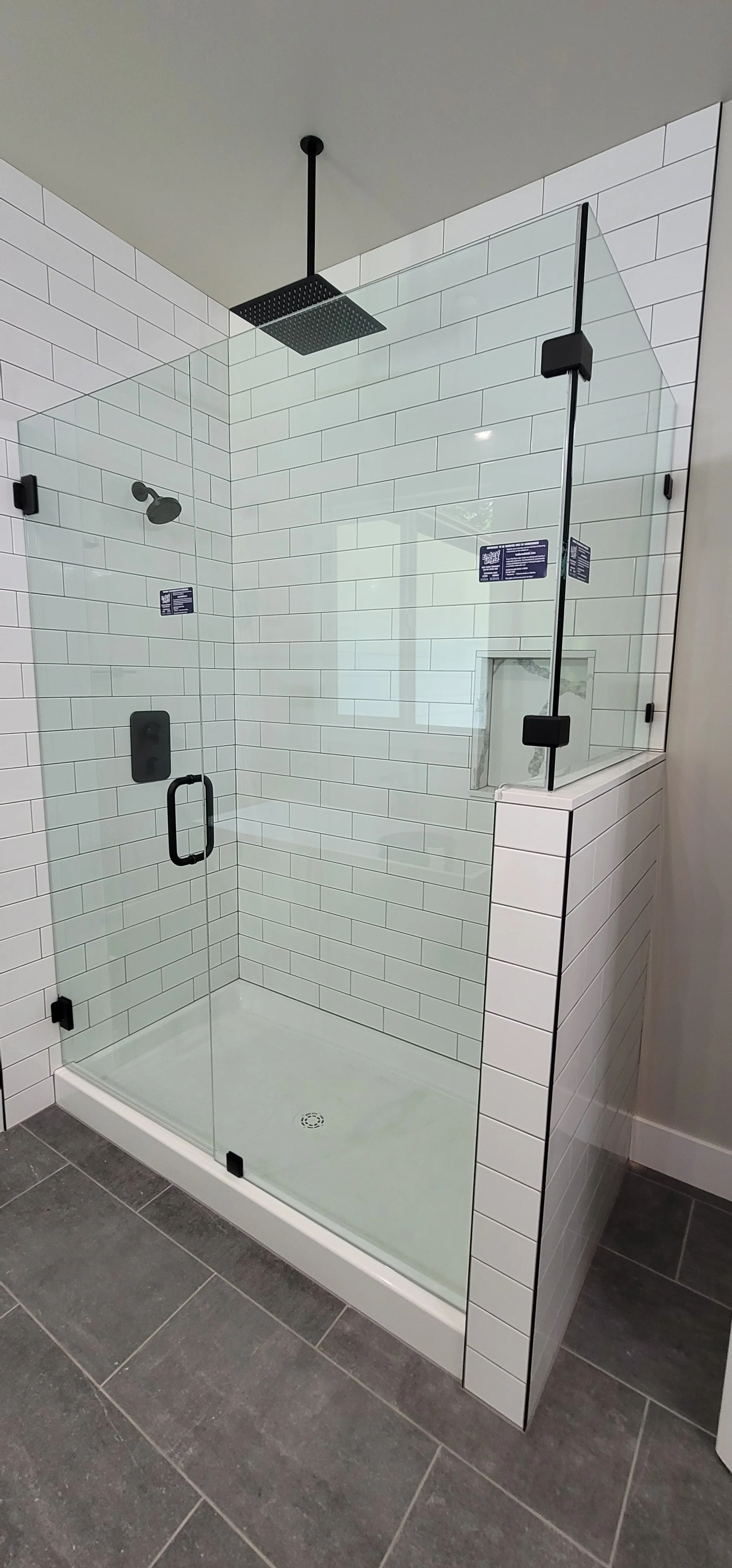 A modern glass-enclosed shower with white subway tile walls, a black rain shower head, a black wall-mounted shower control, and a small built-in niche for toiletries. The shower has a black handle on the door and is installed on a dark tiled bathroom floor.