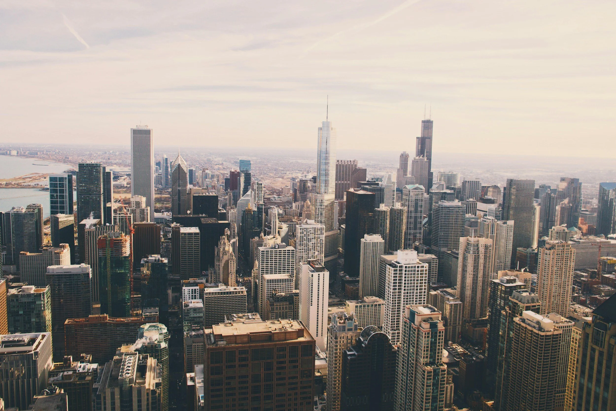 Aerial view of a densely packed city skyline with numerous high-rise buildings, including the John Hancock Center and Willis Tower, with the Chicago River and Lake Michigan visible in the distance.