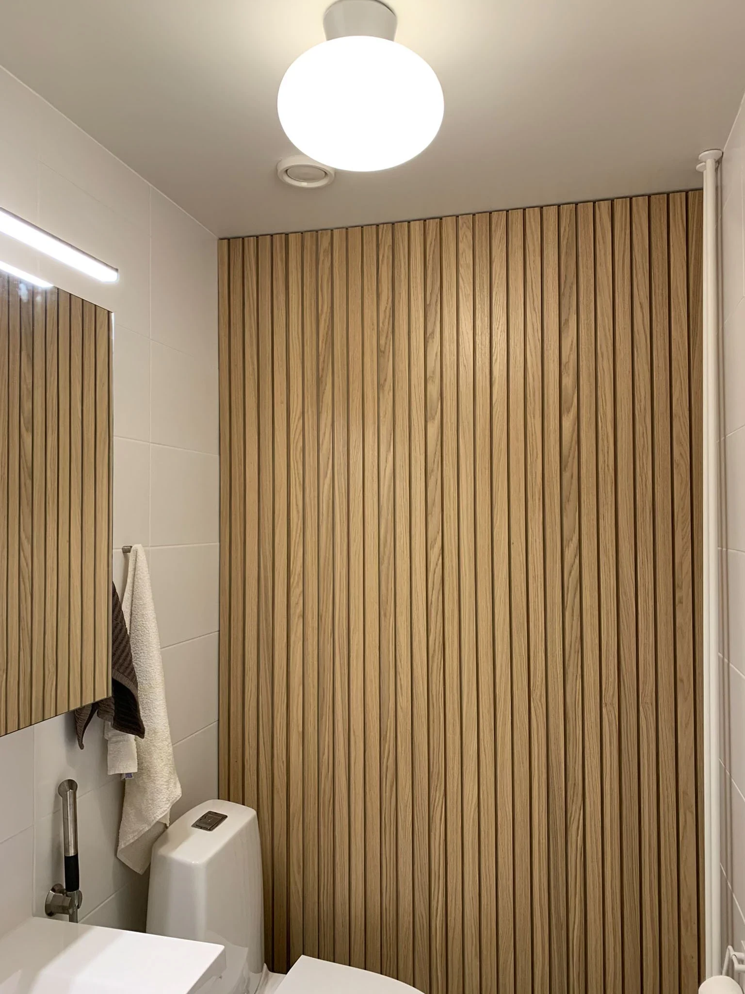Kuvassa selkeä wc-tila, jossa on pyöreä kattovalaisin, puupanelointi osassa seinää, wc-istuin ja peili altaan yläpuolella.