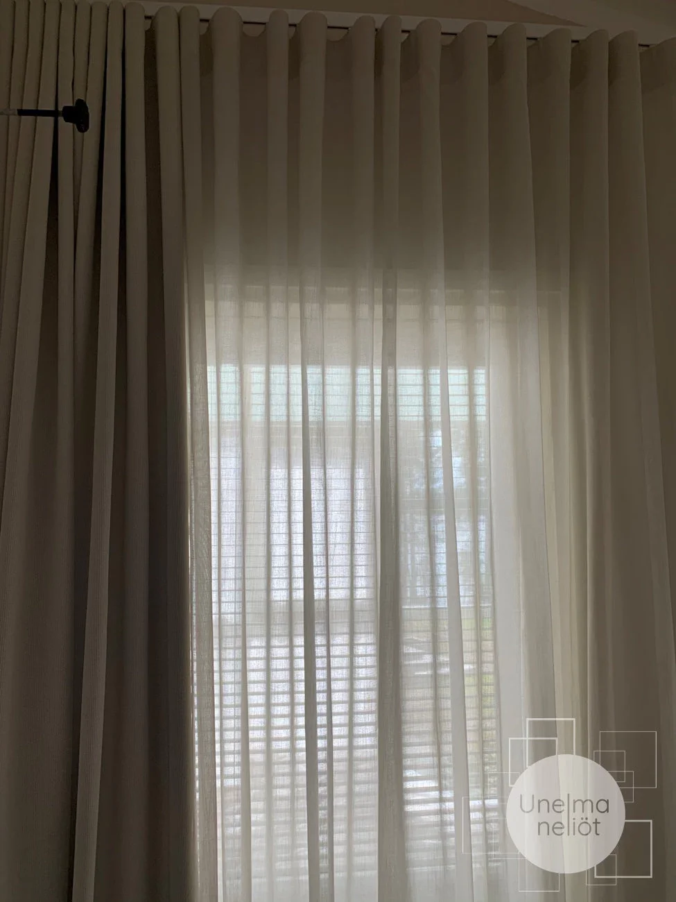 Harmaa verho ja ohuet valkoiset verhot ikkunassa, näytetään pihalle ja rakennuksia.