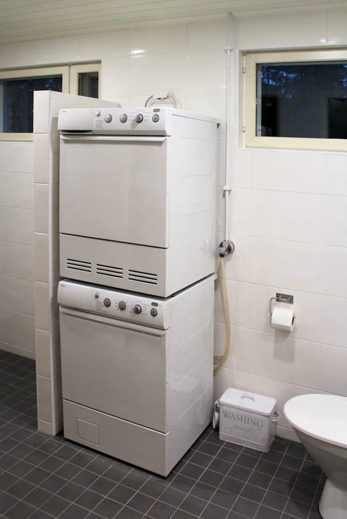 Pesukone ja kuivausrumpu samassa huoneessa päällekkäin, wc-istuin ja paperiteline.