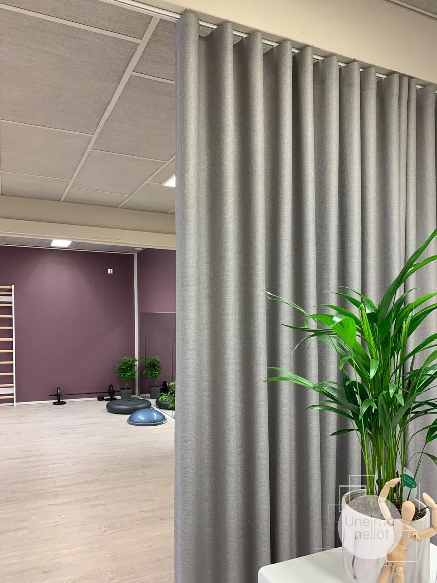 Sisätiloissa oleva tila, jossa on beige verho ja vihreä kasvi. Taustalla näkyy liikuntalattia, seinä ja osa liikuntavälineistä.