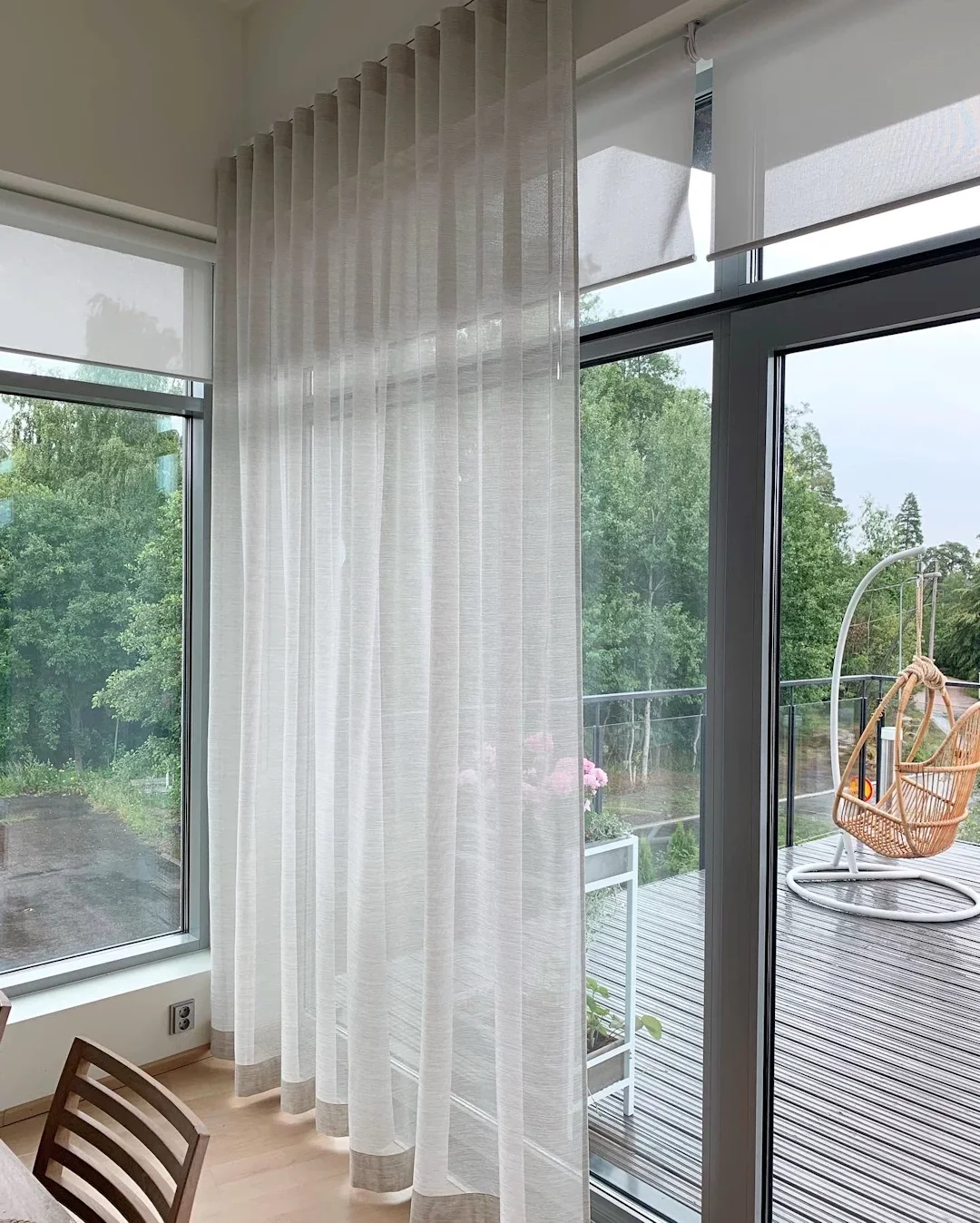 Kuvassa on kerroksinen verhokappa ja lasiovet, jotka avautuvat parvekkeelle. Parvekkeella on rottinki-kelosänky ja kasvilautoja vieressä.