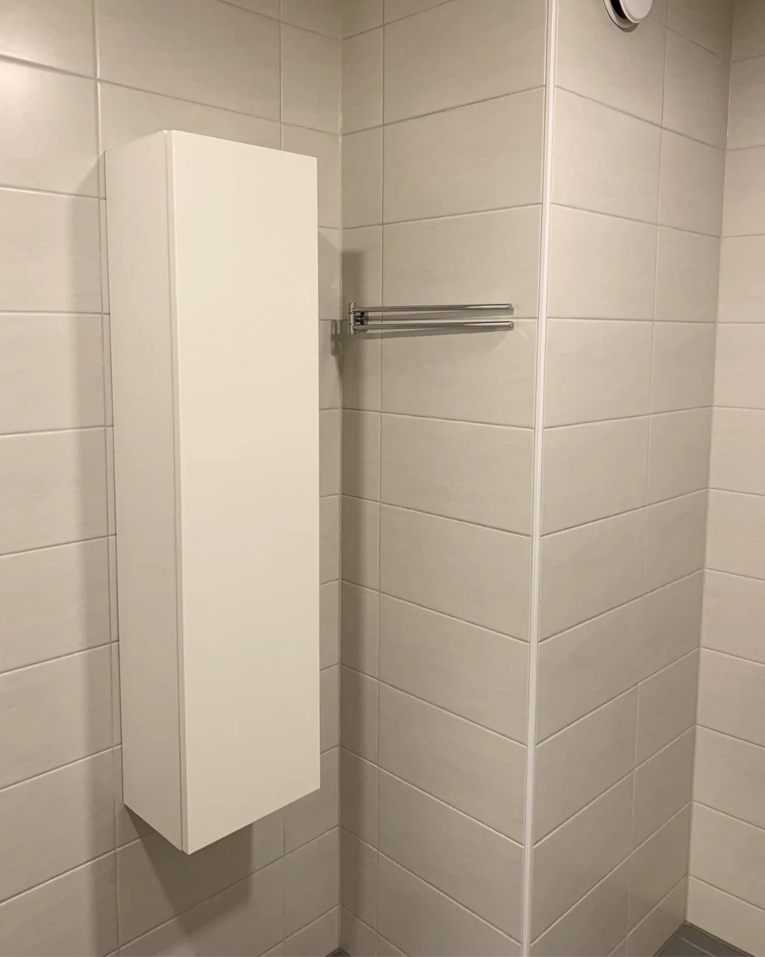 Valkoinen kaappi ja teräksinen pyyheteline kylpyhuoneen seinällä, vaaleat laatat.
