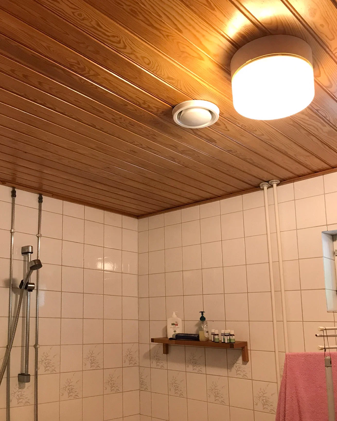 Kylpyhuone, jossa puunväriset kattopaneelit, vaaleat laatat, suihku ja seinähylly shampolle.