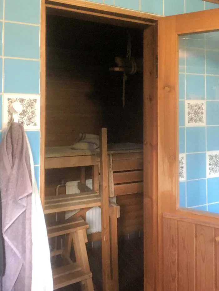 Kuva saunaosastosta, jossa on puisia istuimia ja lämpömittari seinällä, sekä osittain näkyvä kylpyhuoneen seinä, jossa on turkoosikuvioiset laatat.
