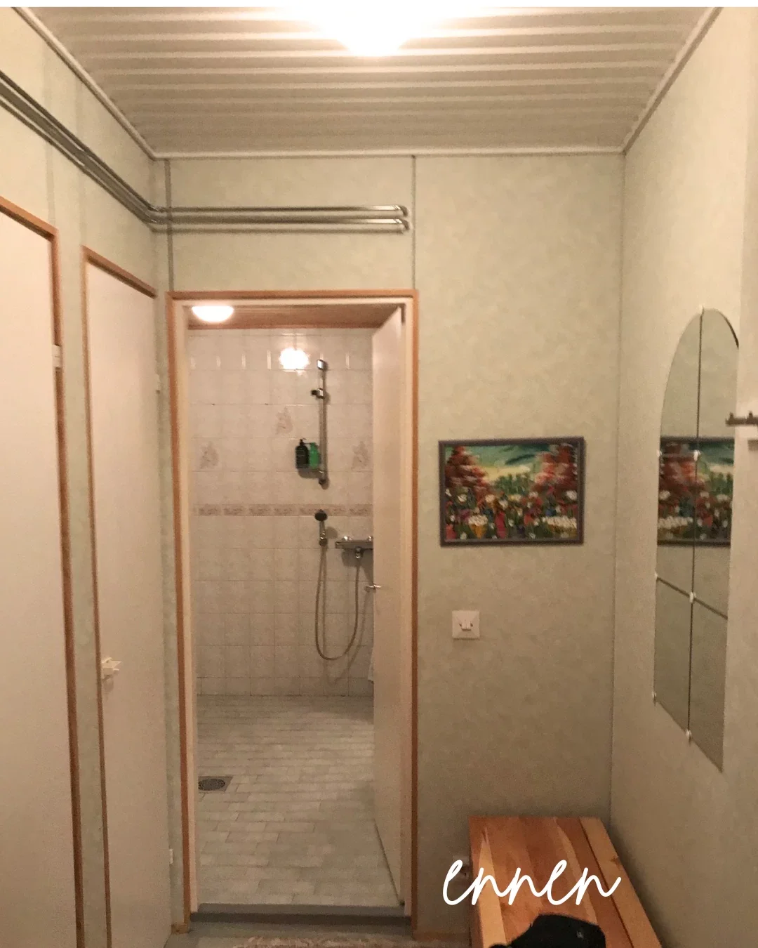 Kuvassa on pieni kylpyhuone, jossa on suihku. Se on erillinen tila, näkyy valkoinen laattaseinä, suihkuverho ja suihkupää. Seinällä on kaksi maalausta, ja vasemmalla on melko pieni peili. Edessä on pieni puupenkki, ja osittain näkyy otsa- ja kasvojen pää. Ympärillä on valkoiset ja beige-väriset seinät ja katto.