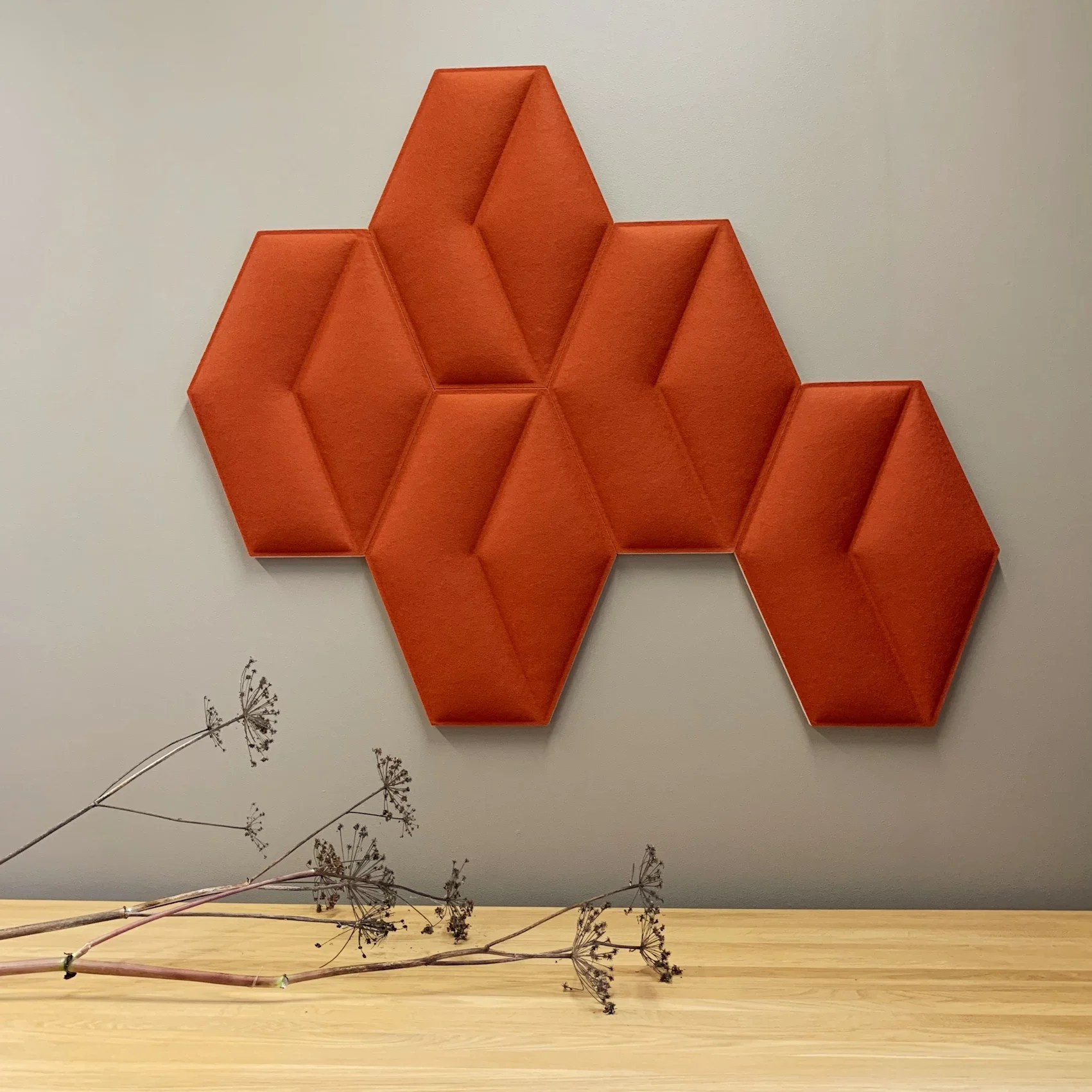 Oranssin värinen geometrinen akustiikkapaneelikokonaisuus seinällä. Kuivattu kasvi asetettuna puiselle pöydälle, taustalla valoisa ja vaalea seinä.