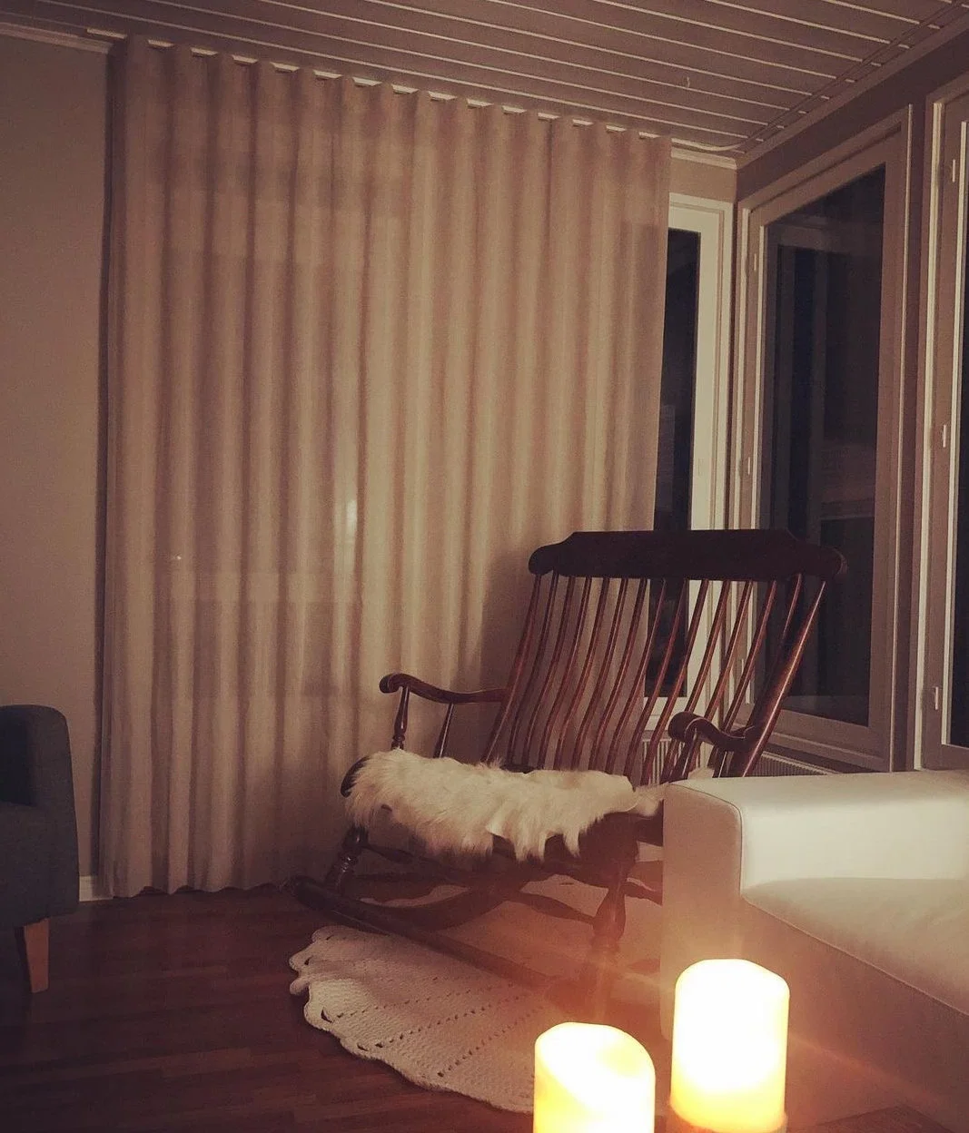Olohuoneen nurkkaus, jossa on keinutuoli, kynttilöitä ja verhot.
